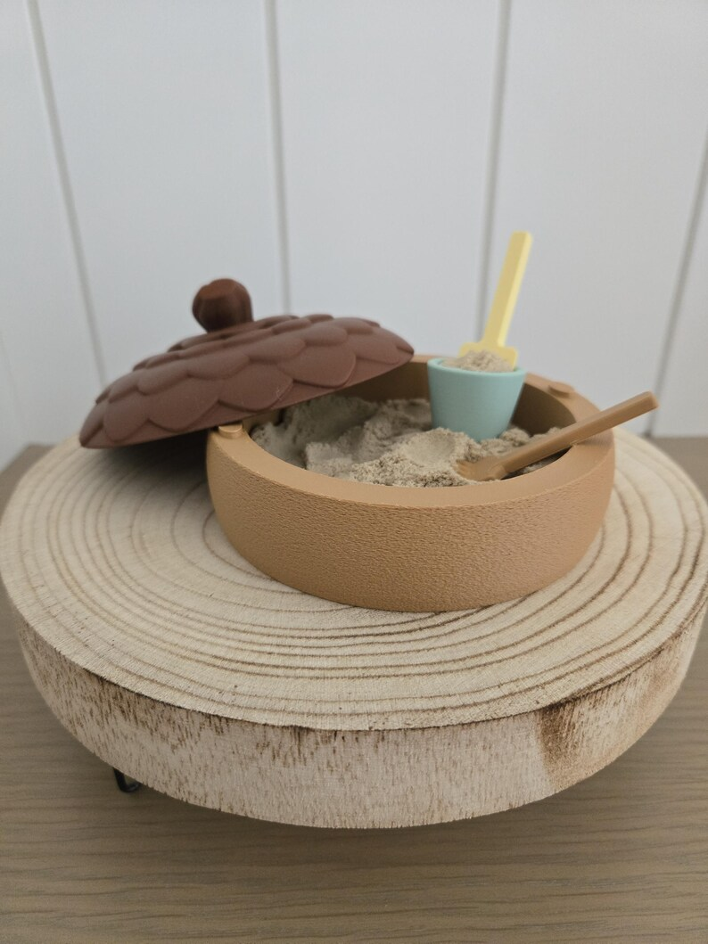 An acorn sandbox with a brown textured lid, sand, a small bucket, and miniature tools.
