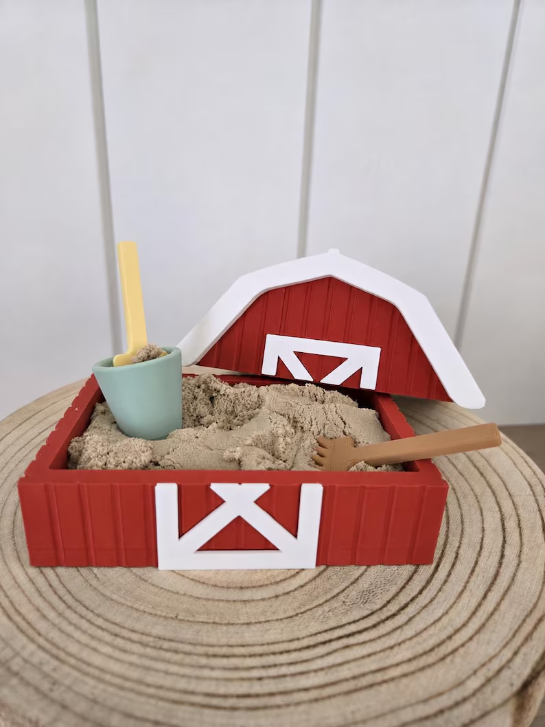 A red barn sandbox with white trim, sand, a small bucket, and miniature tools.