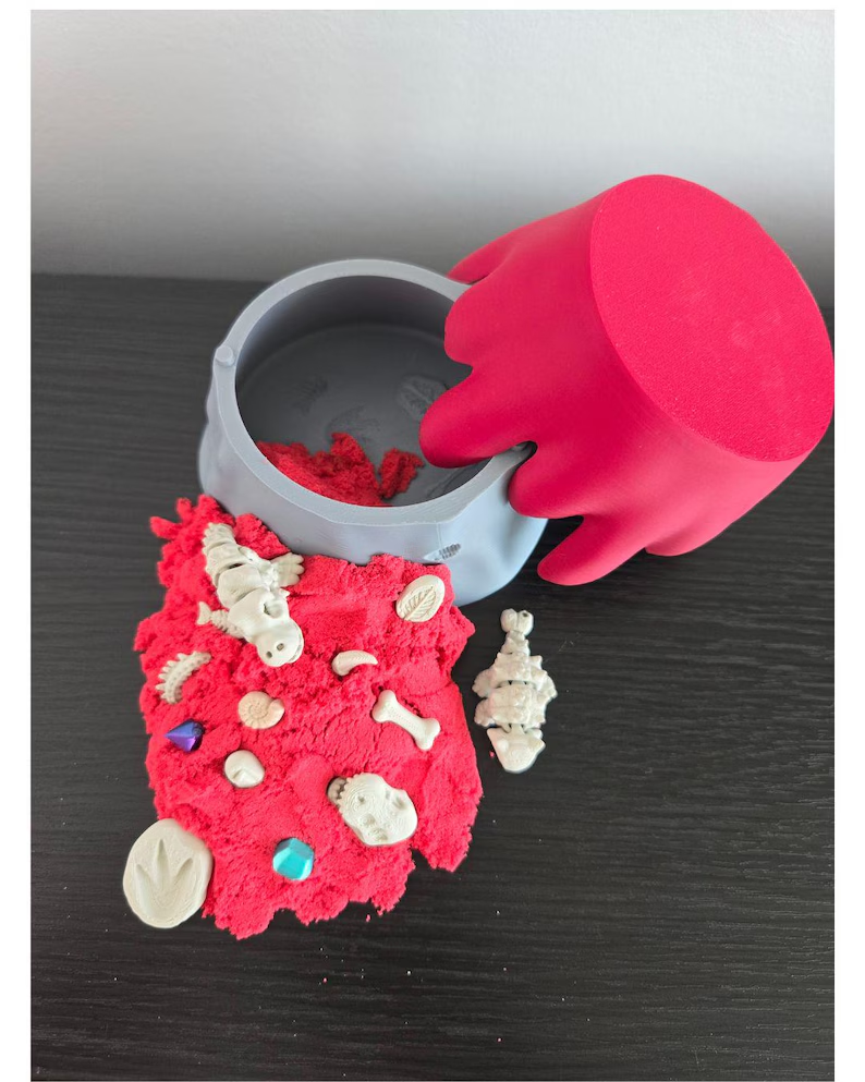 An open gray volcano-shaped container with a red lid, red sand, and miniature dinosaur fossils.