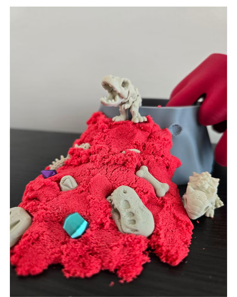Miniature dinosaur fossils and colorful pieces arranged in red sand beside a volcano-shaped container.