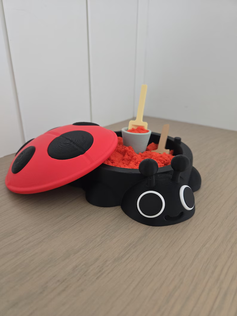 A red and black ladybug sandbox with its spotted lid open, red sand, and miniature tools.