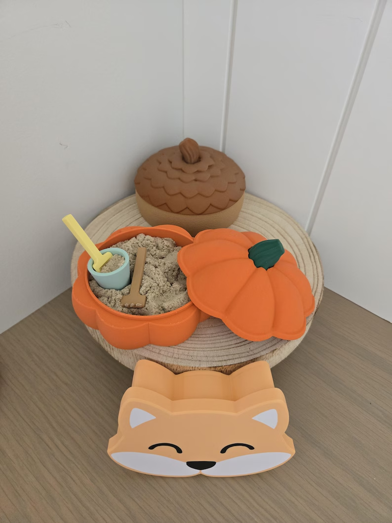 An open orange pumpkin sandbox with sand and miniature tools, with an acorn behind it.