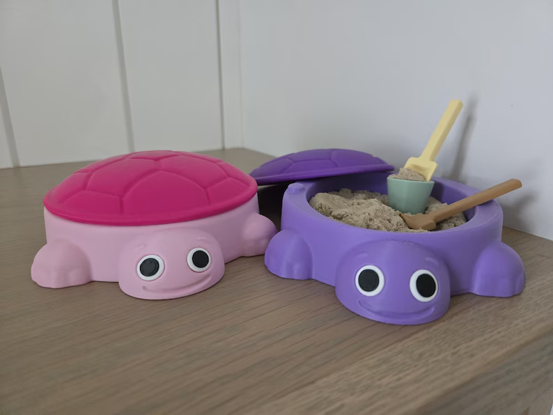 Pink and purple turtle sandboxes, one closed and one open with sand and miniature tools.